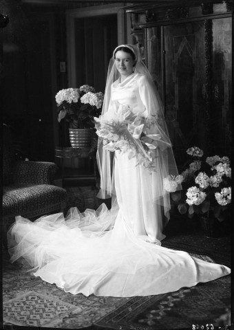 Debout dans un salon une mariée, vêtue de sa longue robe et d'un voile de mousseline, dans ses bras des arums (même cliché que 51 Fi 602) : négatif souple 12,6x17,6 cm.