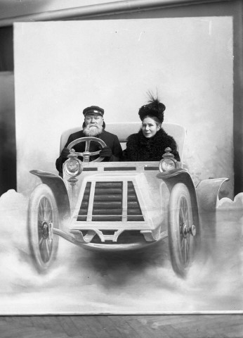 Dans un décor peint sur une toile, un couple en vêtements d'extérieur mime un voyage en automobile : plaque de verre 13x18 cm.