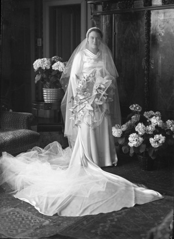 Debout dans un salon une mariée, vêtue de sa longue robe et d'un voile de mousseline, dans ses bras des arums (même cliché que 51 Fi 602) : plaques de verre 13x18 cm.
