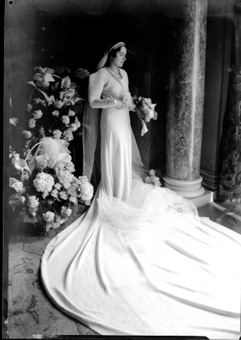 La mariée debout, sa traîne étalée à ses pieds, au milieu des fleurs.
