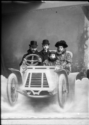 Dans un décor peint sur une toile, une famille en vêtements d'extérieur mime un voyage en automobile : plaque de verre 13x18 cm.