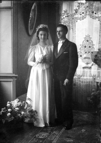 Dans un salon, un couple de mariés non identifié, elle, en robe longue son bouquet à la main, lui, se tient à ses côtés : plaque de verre 13x18 cm, [s.l.].