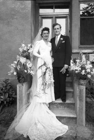 Sur les marches de l'entrée d'une maison. Un couple de mariés pose côte-à-côte. Des corbeilles de fleurs décorent l'entrée.