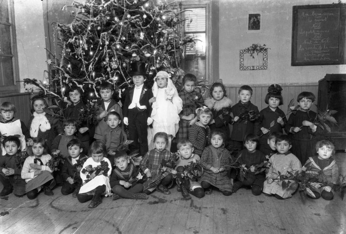 Scénette autour d'un arbre de Noël dans une salle de classe. Les jeunes élèves posent autour d'une petit couple de mariés. Sur le tableau en haut à droite est écrit le programme musical de l'événement : négatif souple 12,6x17,6 cm, [s.l.].
