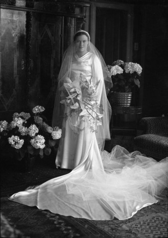 Debout dans un salon une mariée, vêtue de sa longue robe et d'un voile de mousseline, dans ses bras des arums : négatif souple 12,6x17,6 cm.