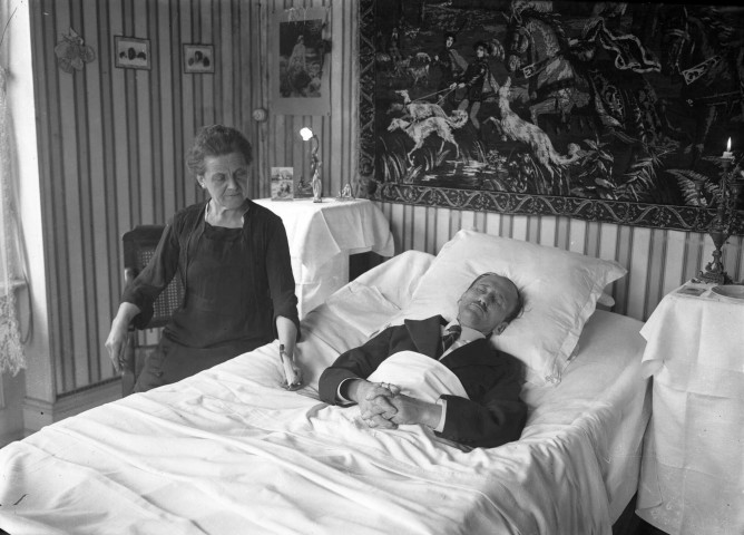 Un homme repose sur son lit de mort les mains jointes ; il est vêtu d'un costume. Une vieille dame assise veille à ses côtés.