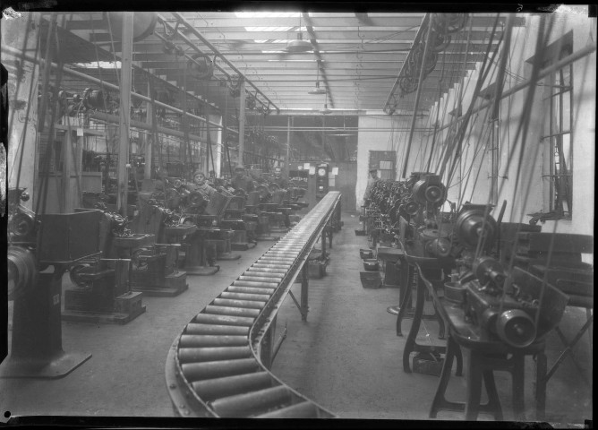 Atelier de machines outils, au premier plan un tapis roulant métallique, des ouvriers.
